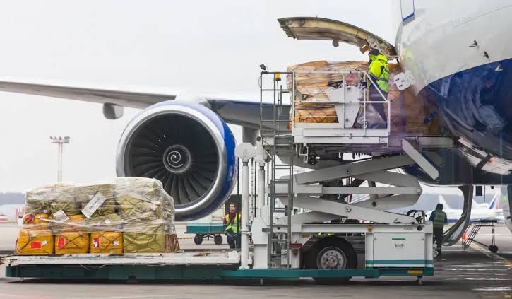 Ground crew loading wrapped cargo pallets into the lower deck of a passenger airplane using a high-lift loader, representing the process of how cargo gets onto a flight.