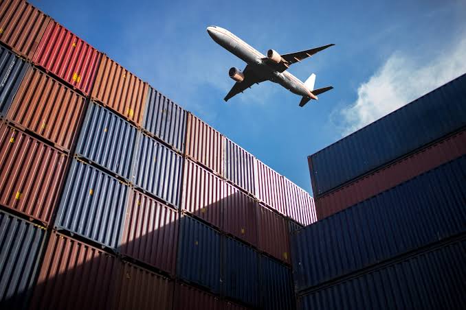 A cargo plane flying over stacked shipping containers against a blue sky, representing the speed and scale of air freight shipments.